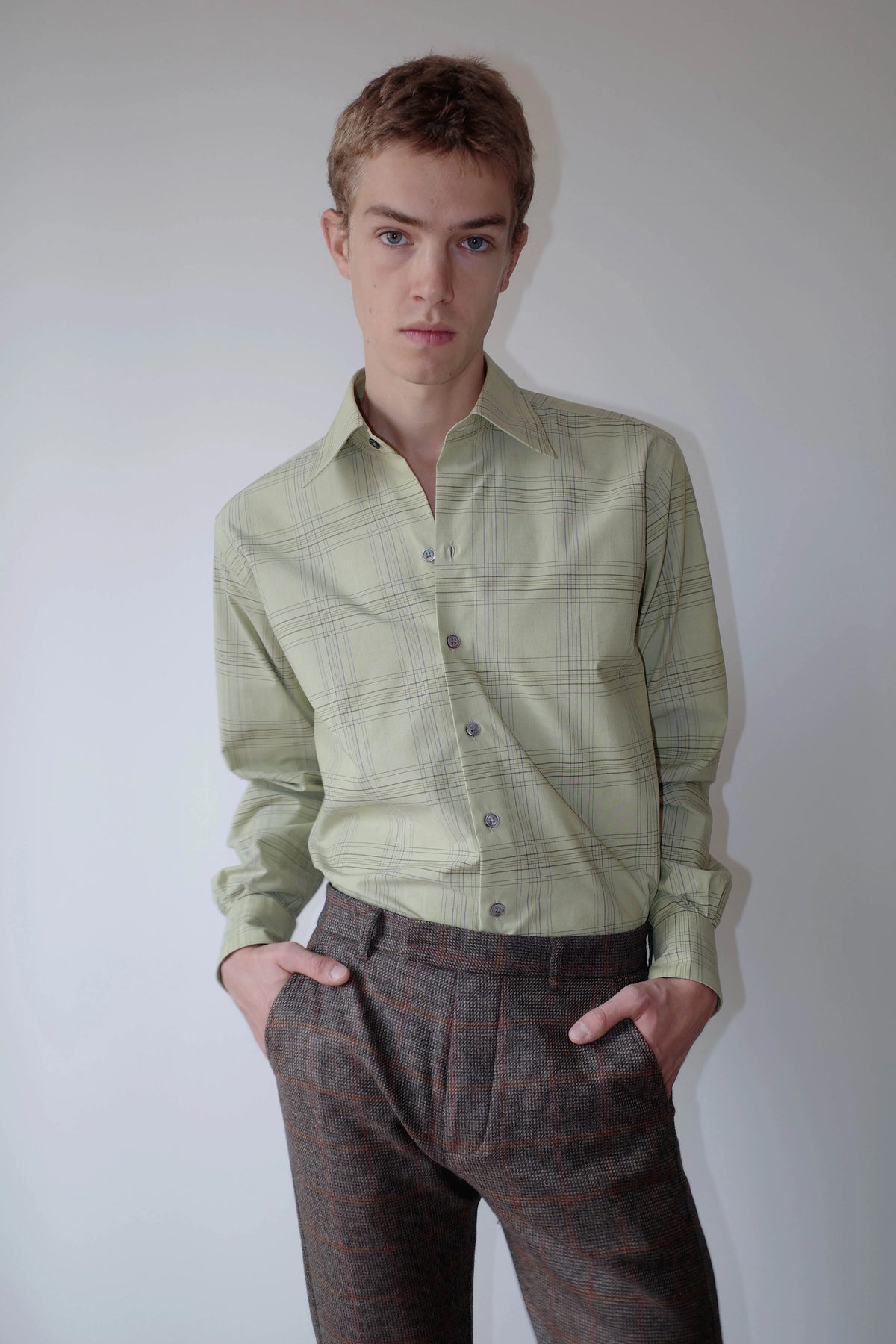 CAMISAS MANOLO（カミサス マノロ）Normal Shirt Pale Green Blue Checksのスタイリング画像。淡いグリーンのチェックシャツをチェック柄のトラウザーにタックインした着用スタイル。自然な生地のドレープと襟元のディテールが特徴。
