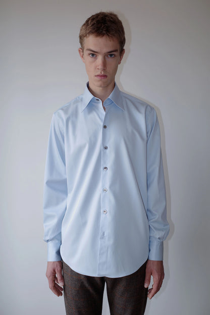 CAMISAS MANOLO（カミサス マノロ）Normal Shirt Light Blue 正面着用画像。淡いライトブルーのコットンツイル生地を使用した長袖シャツ。リラックス感のあるシルエットとラウンドヘムが特徴。