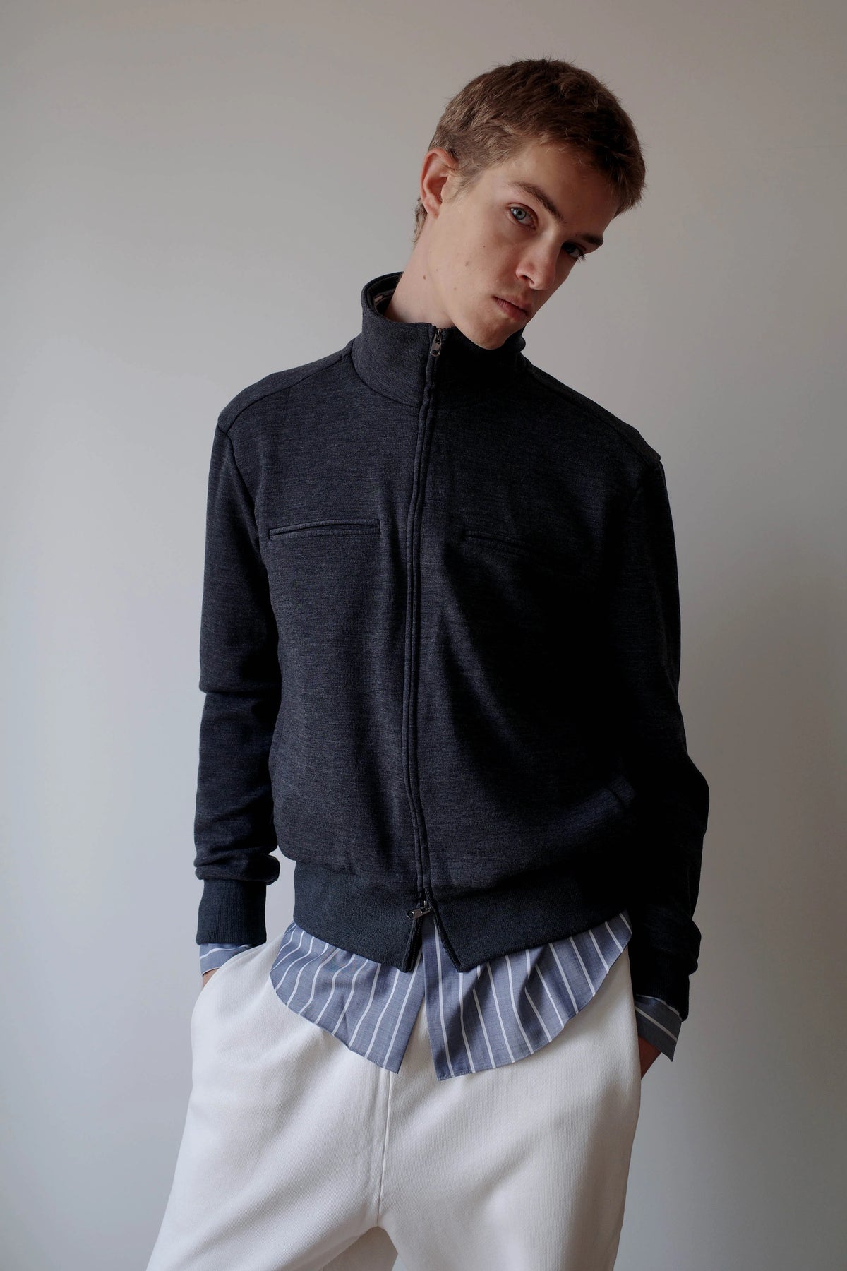 CAMISAS MANOLO（カミサス マノロ）Jersey CM Jacket Greyのスタイリング画像。グレーのジップジャケットにブルーのストライプシャツとホワイトのトラウザーを合わせた着用スタイル。ダブルジップを閉じたスタンドカラーと、裾から覗くシャツのレイヤードが特徴。