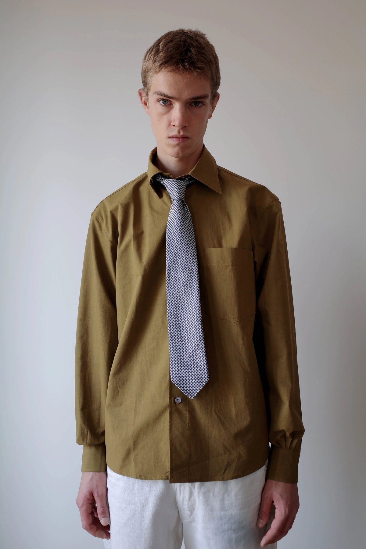 CAMISAS MANOLO（カミサス マノロ）Sobrecamisa Shirt Oliveのスタイリング画像。オリーブカラーの長袖シャツに柄物のネクタイとホワイトのトラウザーを合わせた着用スタイル。オーバーサイズのシルエットと落ち着いた色合いが特徴。