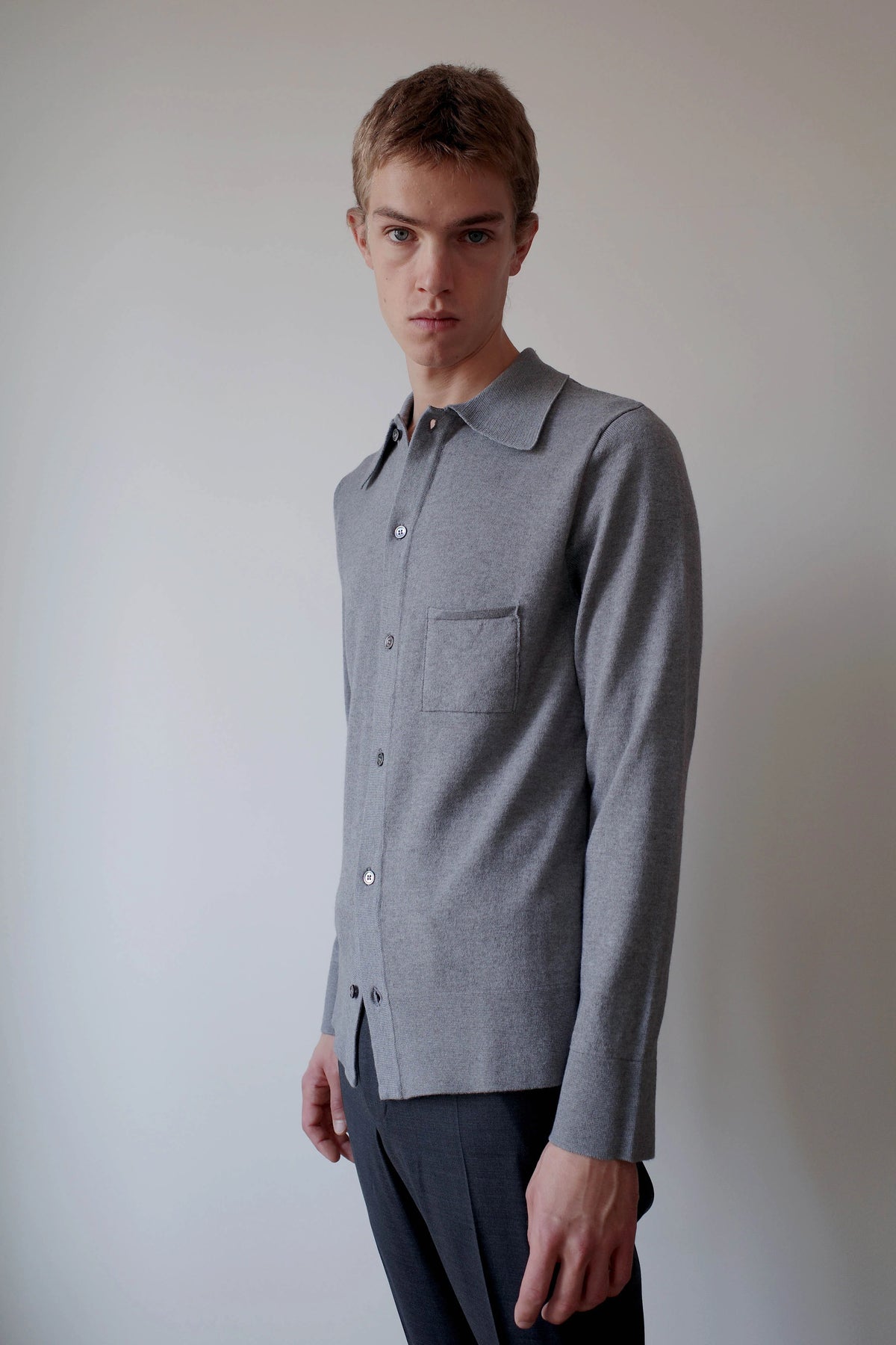 CAMISAS MANOLO（カミサス マノロ）Knitted Shirt Light Greyの斜め前からの着用画像。柔らかなライトグレーのニットシャツで、自然な生地の落ち感とすっきりとしたシルエット、襟元や胸ポケットのディテールが確認できる。