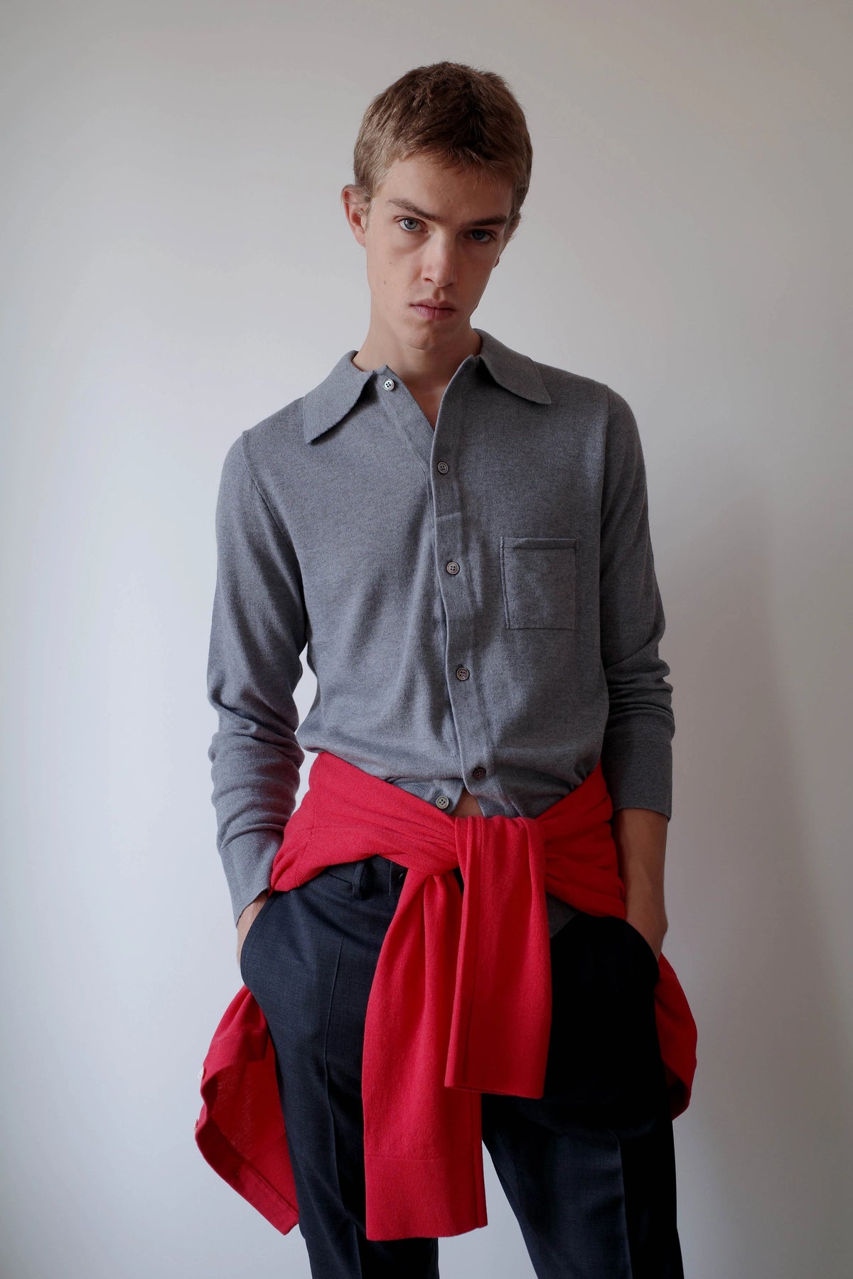 CAMISAS MANOLO（カミサス マノロ）Knitted Shirt Light Greyのスタイリング画像。ライトグレーの長袖ニットシャツにダークカラーのトラウザーを合わせ、鮮やかなレッドのニットを腰に巻いた着用スタイル。袖をまくり、裾をタックインしたコーディネート。
