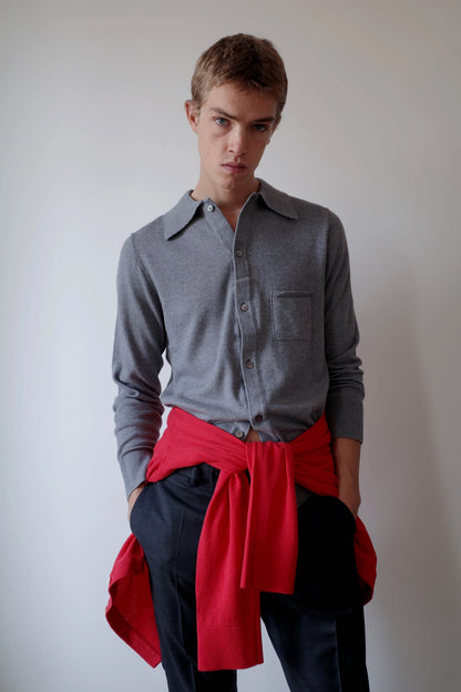 CAMISAS MANOLO（カミサス マノロ）Knitted Shirt Light Greyのスタイリング画像。ライトグレーの長袖ニットシャツにダークカラーのトラウザーを合わせ、鮮やかなレッドのニットを腰に巻いた着用スタイル。袖をまくり、裾をタックインしたコーディネート。