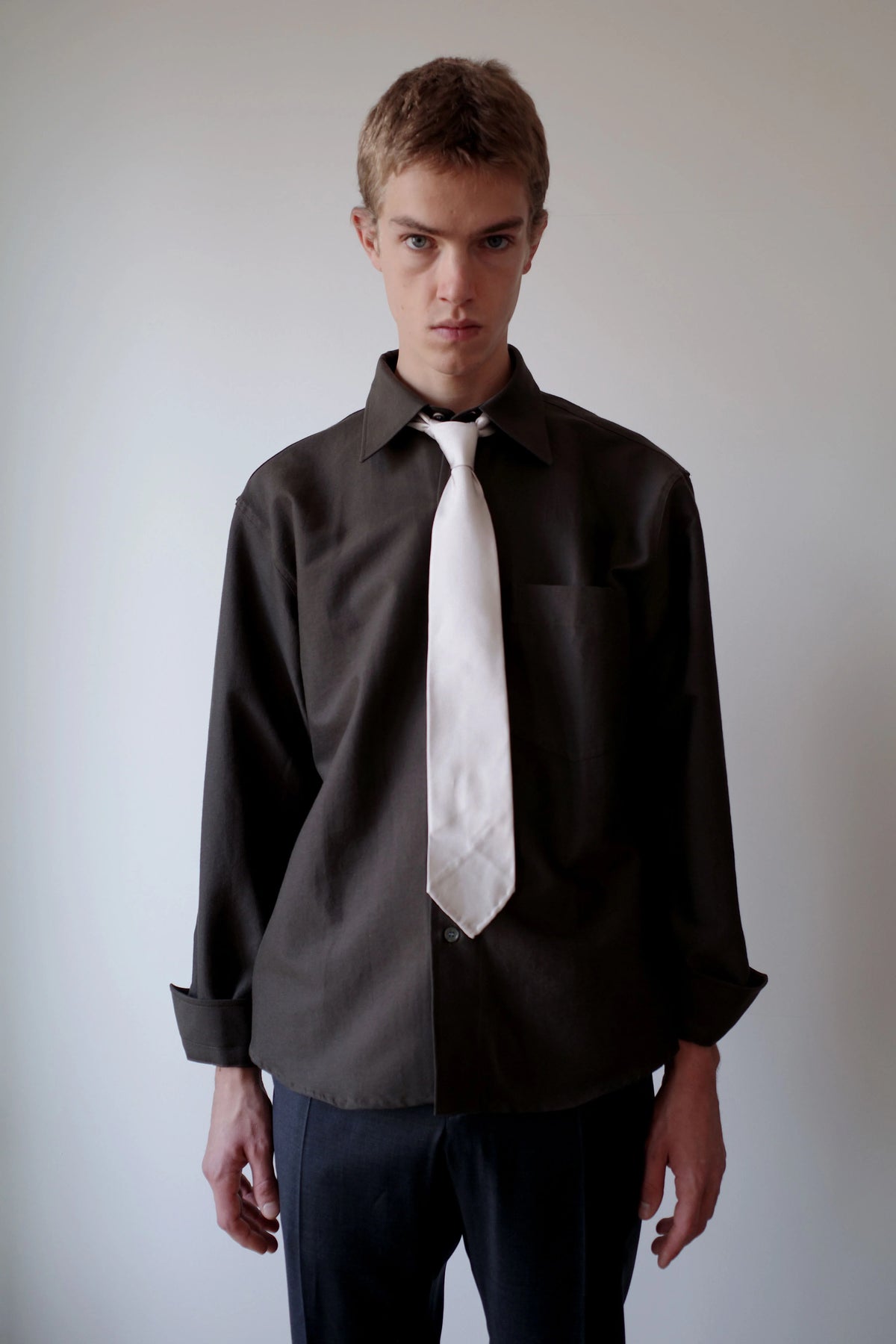 CAMISAS MANOLO（カミサス マノロ）Sobrecamisa Shirt Greenのスタイリング画像。ダークグリーンの長袖シャツにホワイトのネクタイとダークカラーのトラウザーを合わせた着用スタイル。袖口を折り返したリラックス感のあるコーディネート。